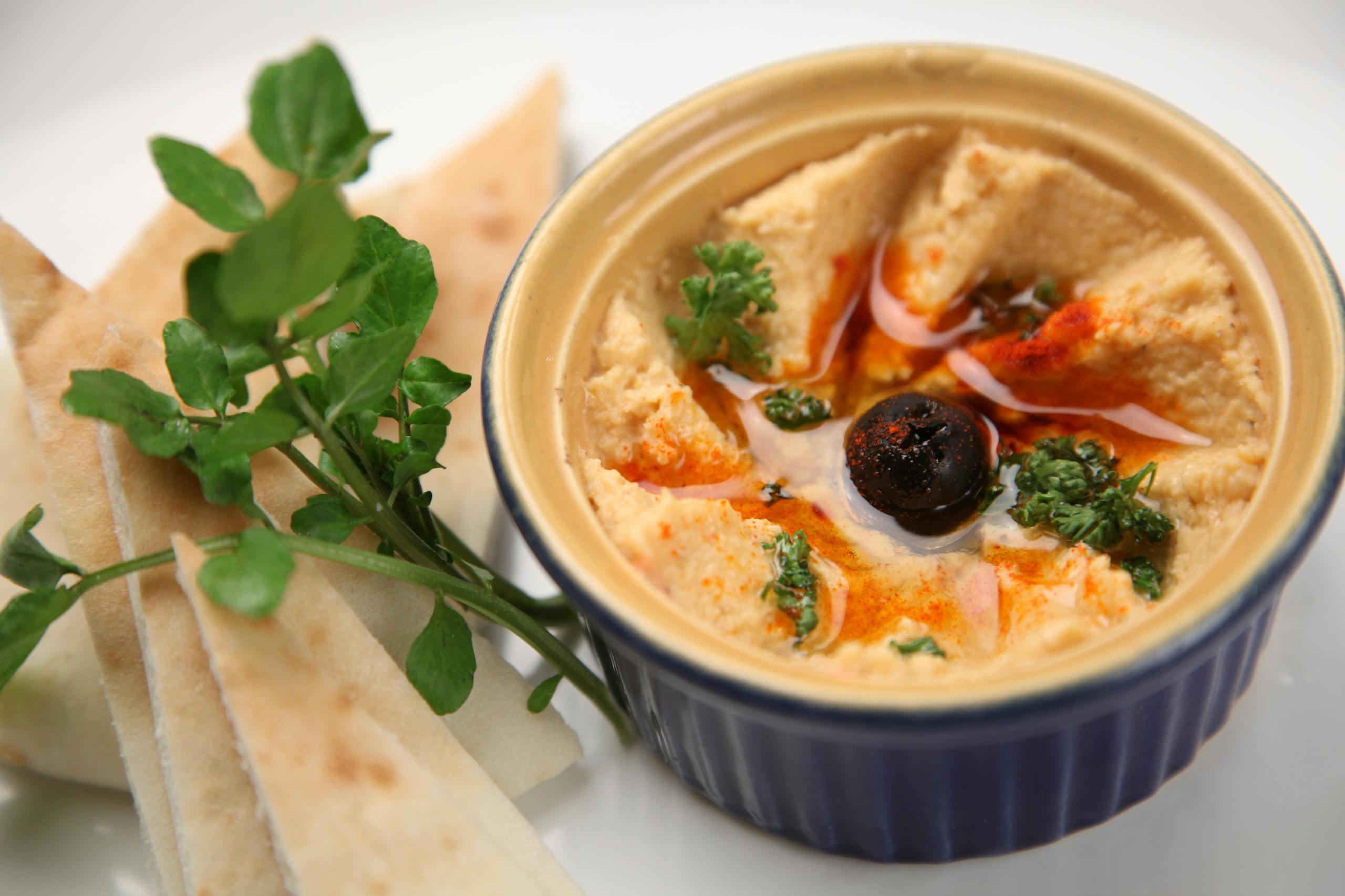 A close-up of creamy homemade hummus with pita slices, garnished with olive and herbs.