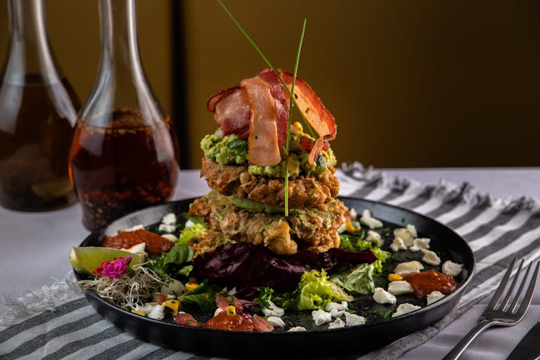 A gourmet salad featuring a tower of bacon and guacamole, served on a black plate with garnishes.