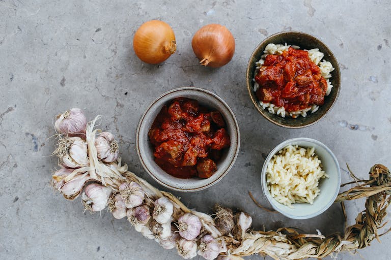 Top view of a rustic meal with garlic, onions, and bowls of rice and stew on a stone surface.