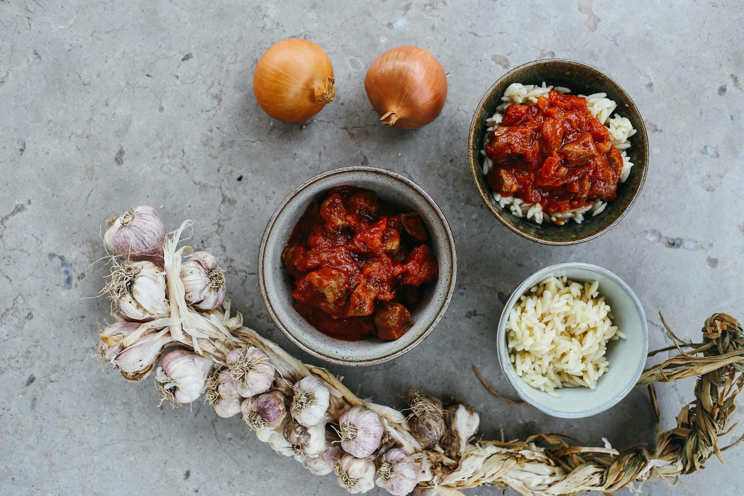 Top view of a rustic meal with garlic, onions, and bowls of rice and stew on a stone surface.